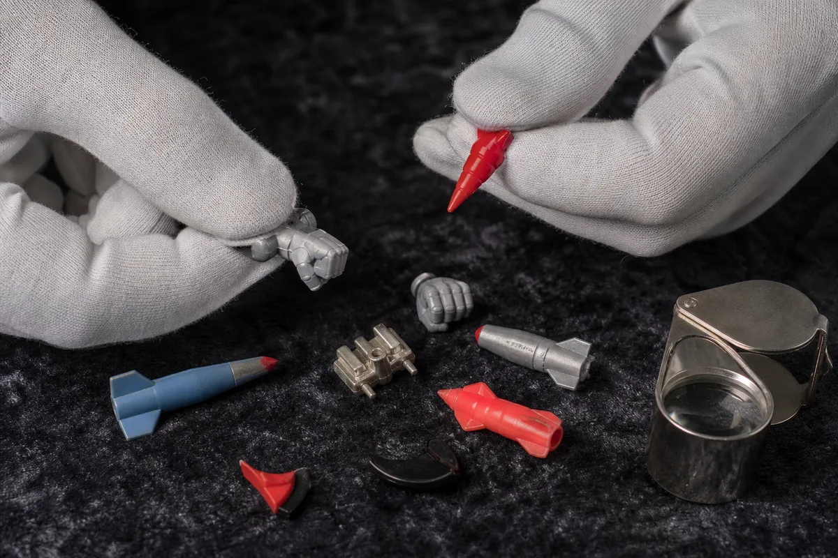 Mains gantées inspectant les accessoires d&rsquo;un jouet robot vintage posé sur un tissu sombre