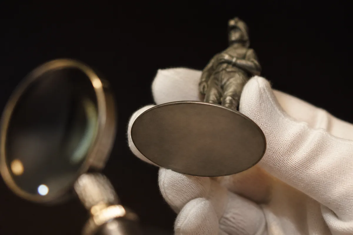 Main gantée présentant le dessous nu du socle d&rsquo;une figurine ancienne sous une loupe